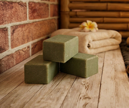 Three green bars of soap on a wooden surface with towels and a flower in the background.