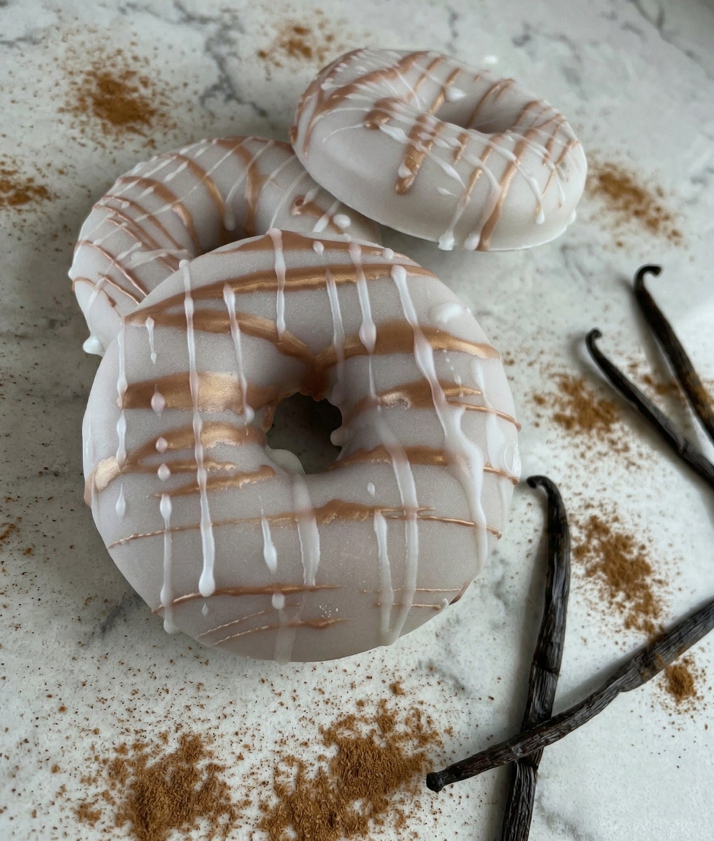 Two white-glazed donuts with brown drizzle on a marble surface with vanilla beans and cinnamon.