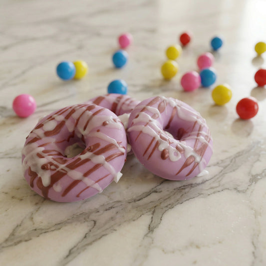 Pink donut-shaped soaps with white drizzle on a marble surface with colorful candies in the background.