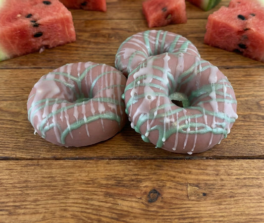 Two pink and green donut-shaped candies on a wooden surface with watermelon slices in the background.