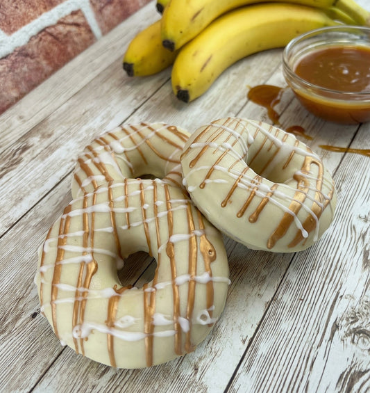 Two donuts with caramel drizzle on a wooden surface with bananas and a bowl of caramel in the background.
