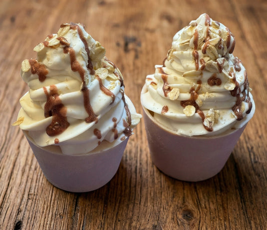 Two soap cupcakes with whipped cream, oatmeal, and caramel drizzle on a wooden surface.
